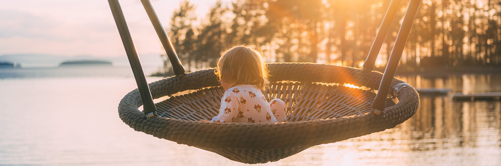 A child swings on a basket swing, looking over to the lake in the background at sunset.